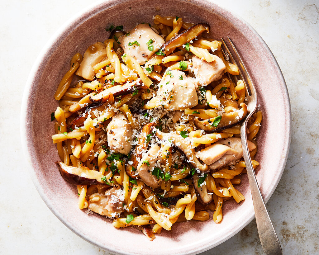 A bowl of pasta, chicken and mushrooms, garnished with Parmesan and parsley. A fork is in the bowl.