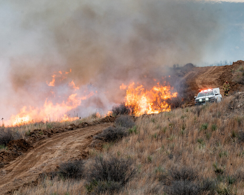 An emergency vehicle and a firefighter stand next to a line of flaming grass.