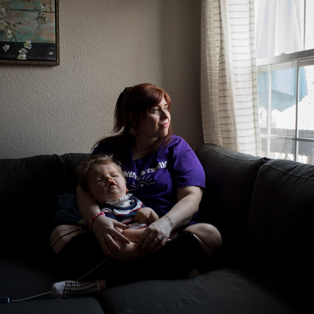 A portrait of Kacey Poynter, who sits on a gray couch and holds her son, Sonny, by a window. A tracheotomy tube is inserted in his neck with a cord leading from it resting on the couch next to them.