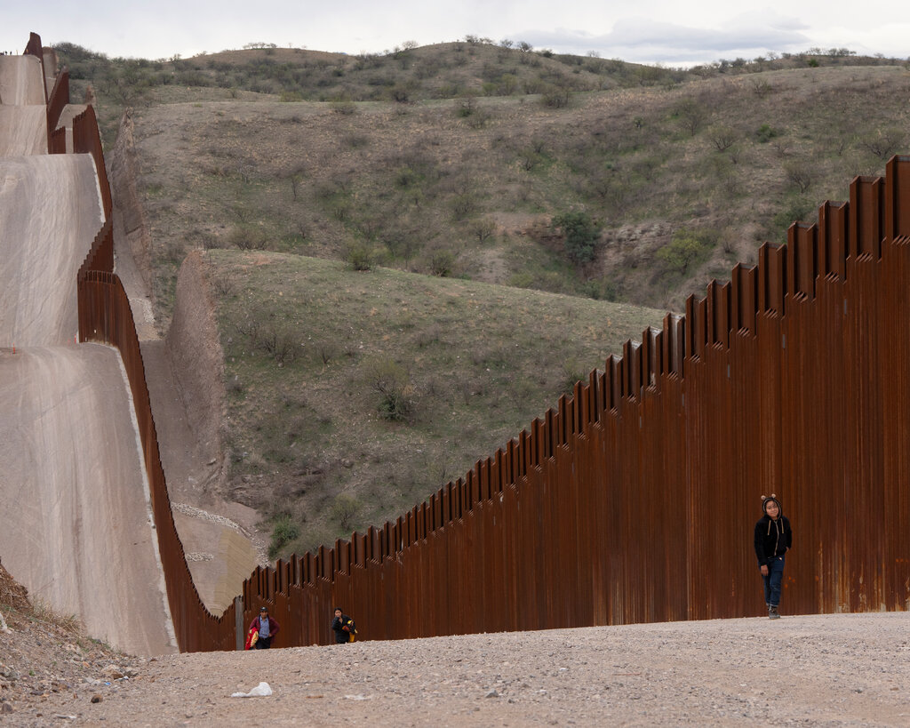 Helen Ramajo, wearing a fuzzy bear-eared hoody is dwarfed by a long, rust-colored barrier, with rolling hills in the background. Several yards behind her are two adults walking.