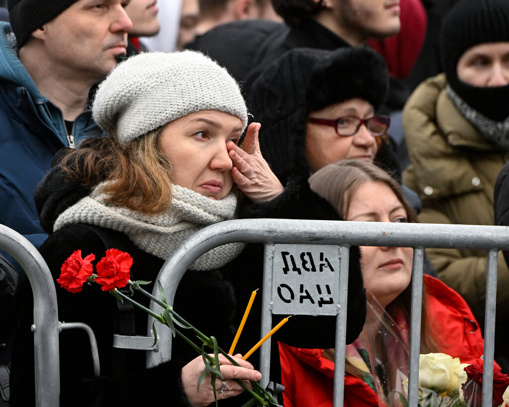 A women in gray hat and scarf, holding red flowers, wipes her eye. Other mourners are behind her.