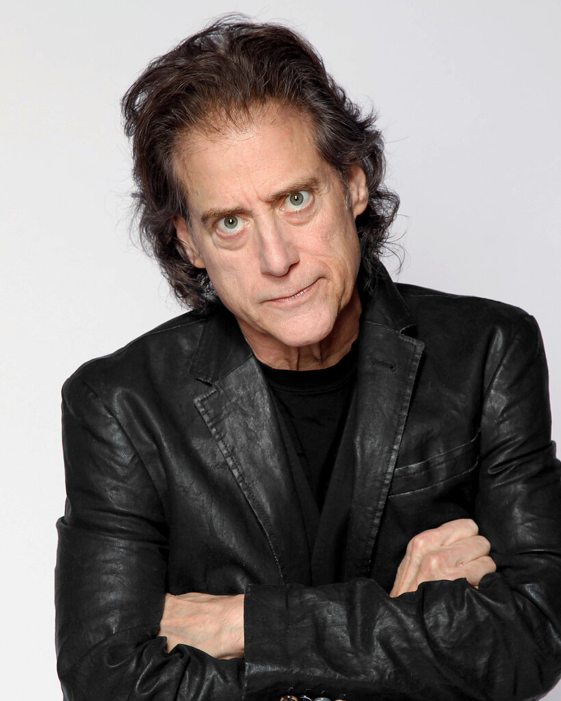 Richard Lewis, an intense-looking dark-haired man wearing a black leather jacket over a black T-shirt with his arms crossed.