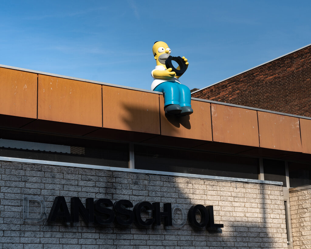 A figure of Homer Simpson sits on top of a school building, eating a large letter O. On a sign below, the letter O is missing from the word “school.”