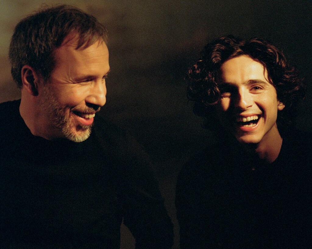 In a shot of two people laughing, Denis Villeneuve is looking at Timothée Chalamet, who’s looking in the camera’s direction. They’re both wearing dark clothes and are pictured from the chest up.