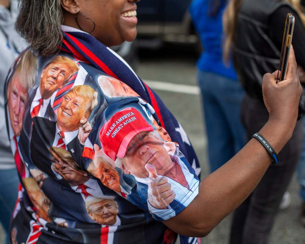 A woman in a shirt covered with Donald Trump’s face holds up her phone. 