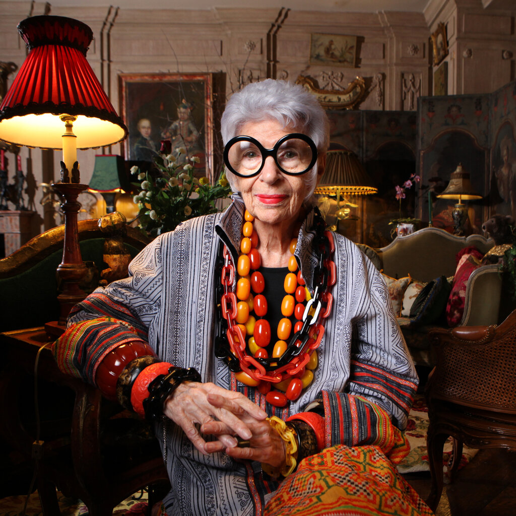 Ms. Apfel sitting in her apartment living room wearing a multicolored outfit with strings of oversize beads around her neck and thick bracelets of many colors on her wrists. The room behind her is crowded with furniture, plants, lamps and many other furnishings.