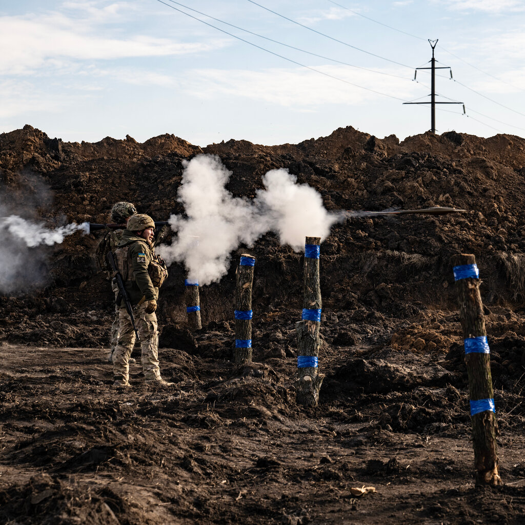Two soldiers stand on bare earth in front of a berm, with poles embedded in the ground and puffs of smoke in the air nearby.