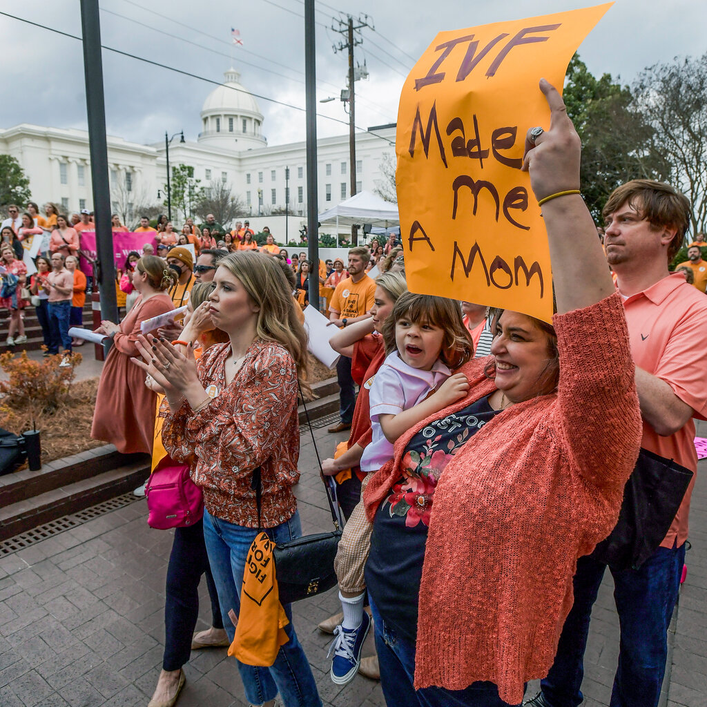 A crowd of protesters, including a woman holding a child and a sign reading “IVF made me a mom,” gathers in front of the Alabama State Capitol.