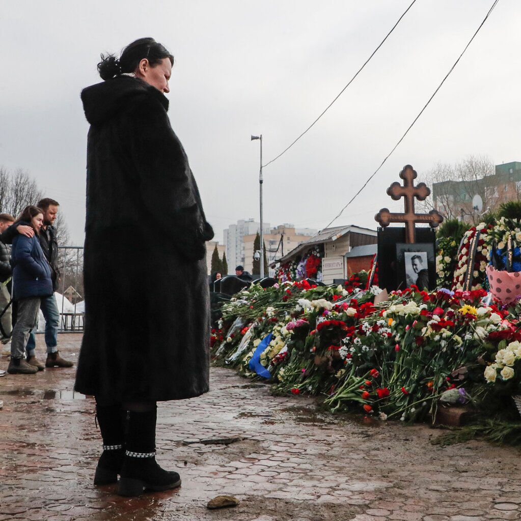 A woman standing by a large pile of flowers, and an image of Aleksei Navalny beneath a Russian Orthodox cross.