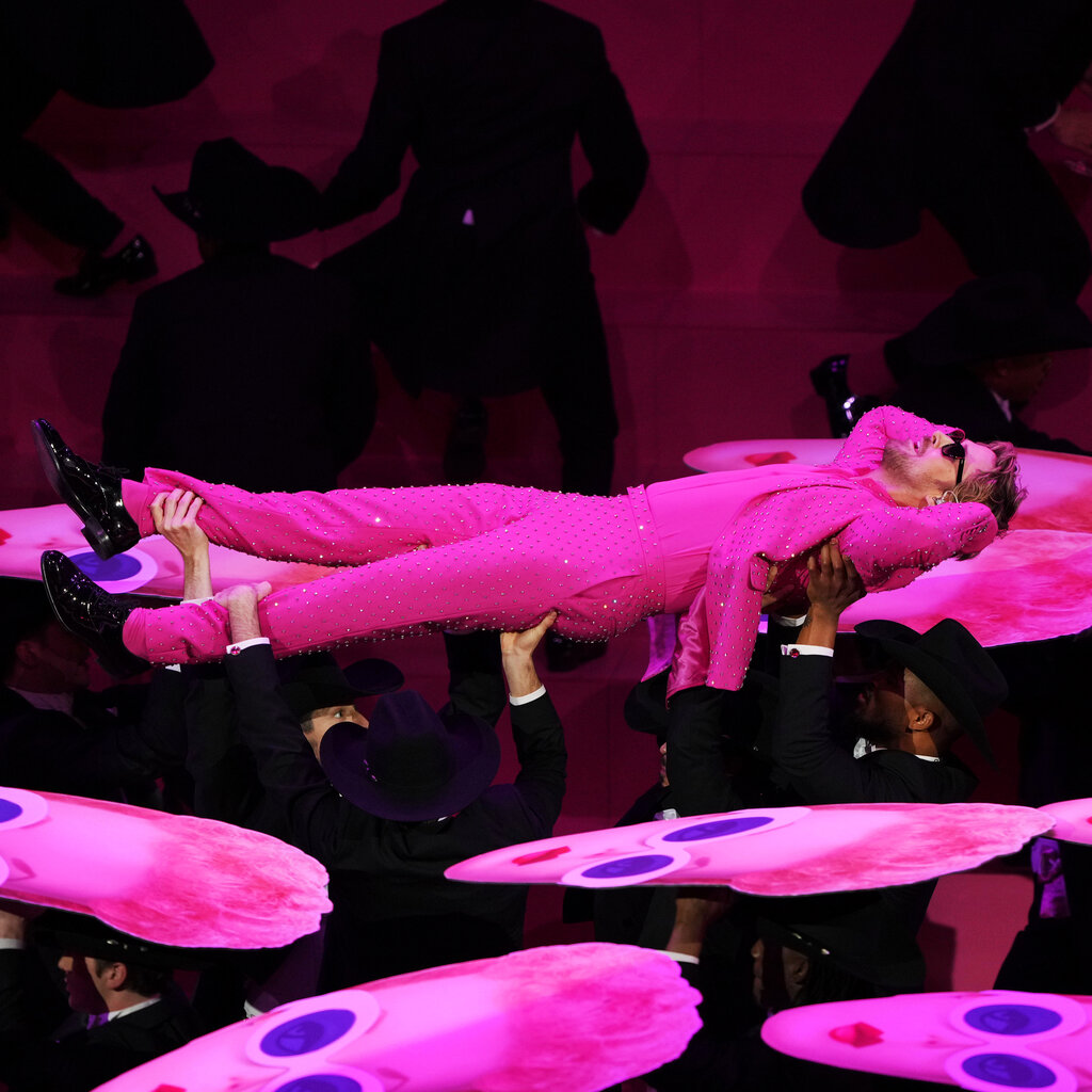Ryan Gosling wearing a pink bedazzled suit and sunglasses, being held up horizontally by men in black suits. 