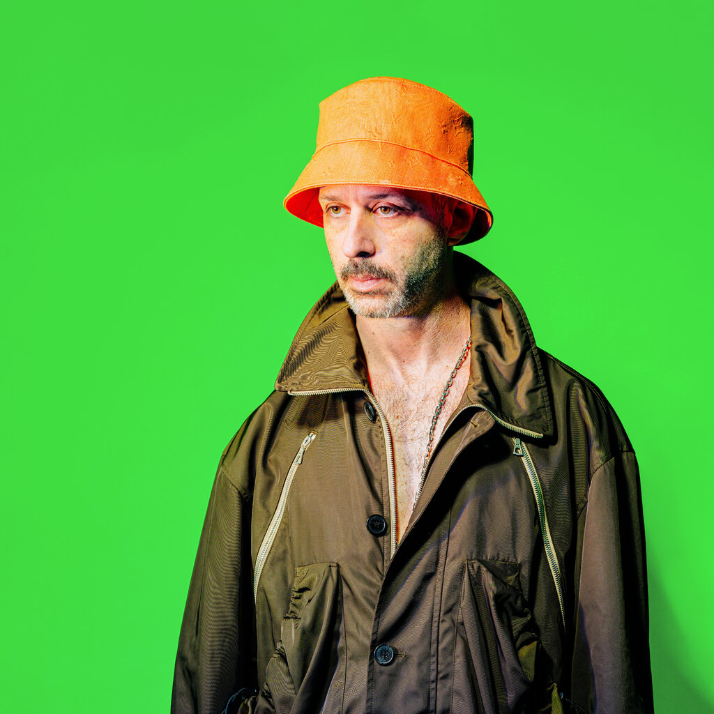 Jeremy Strong wears an orange hat and green jacket against a green screen backdrop. 