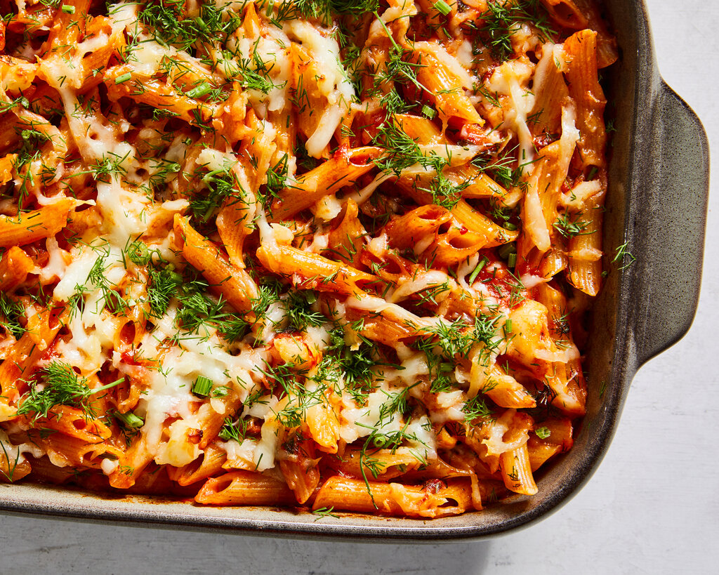An overhead image of a baking dish filled with tube pasta coated in a red sauce and finished with cheese and herbs.