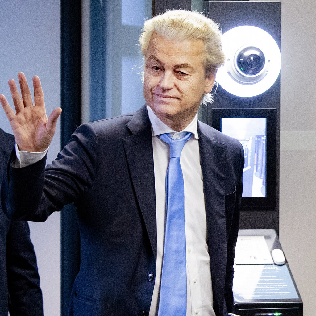 Geert Wilders, in a suit and blue tie, holds up a hand in greeting.