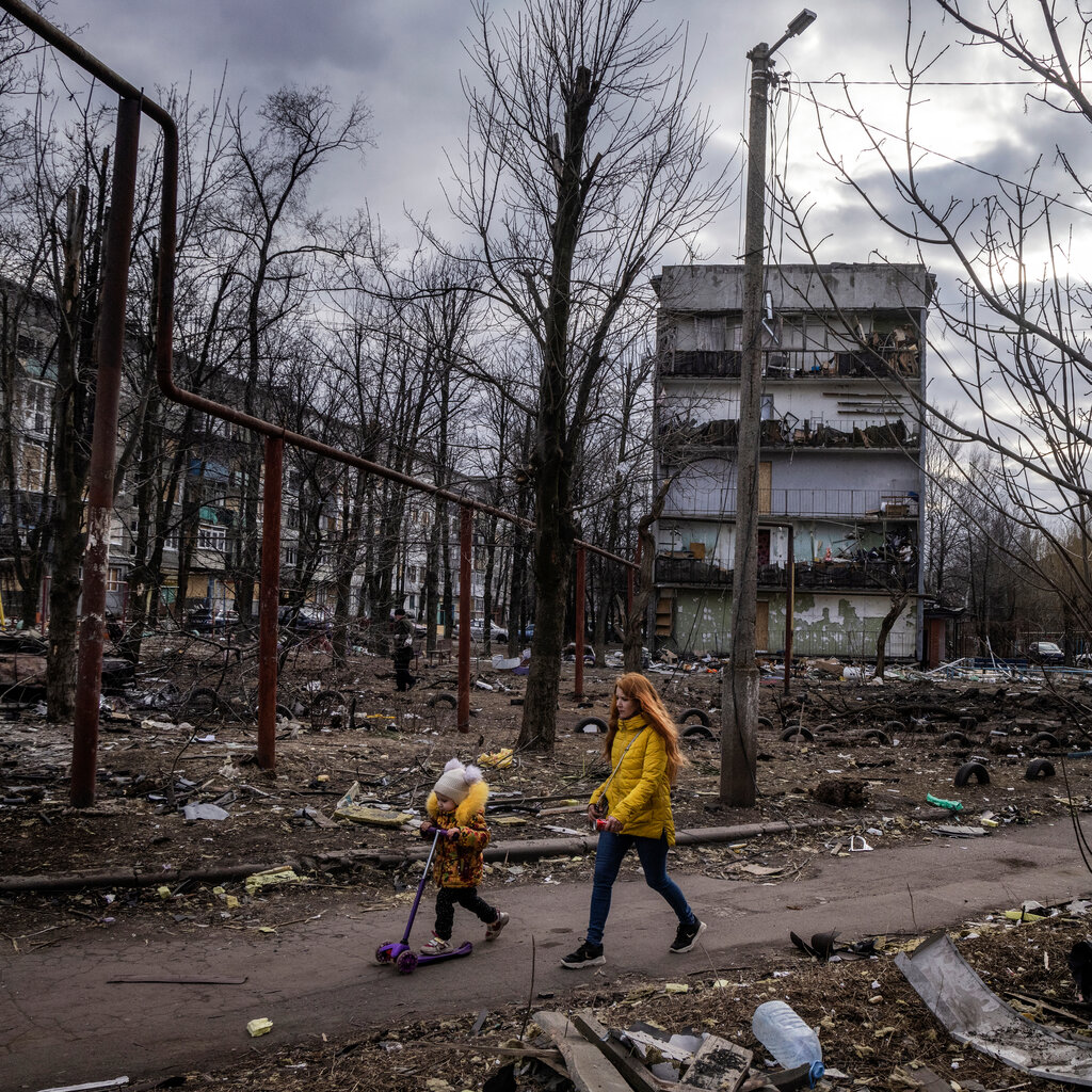 A woman and a child walk through a destroyed neighborhood and damaged apartment buildings.