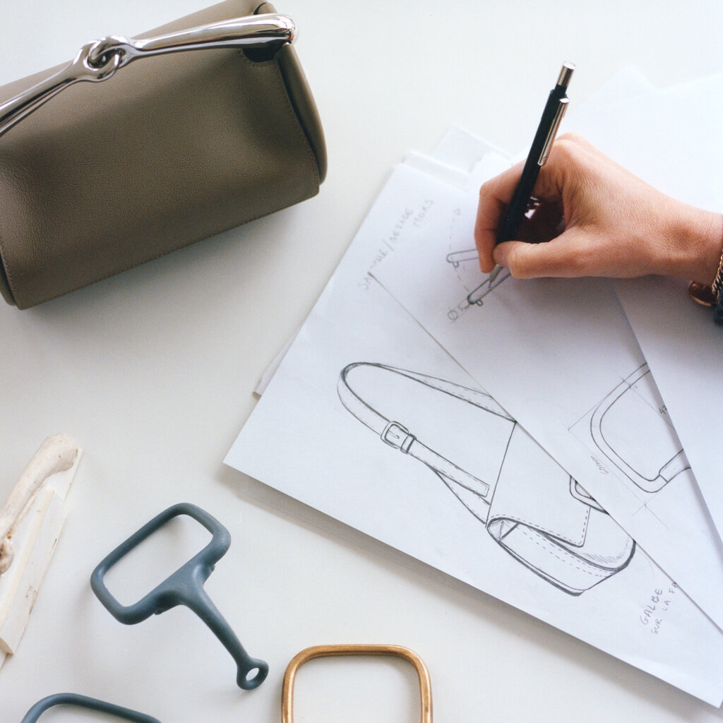 A hand draws purse designs on paper beside a beige handbag.