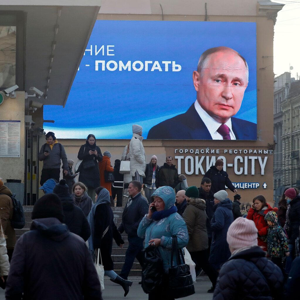 People walk along a street under a billboard with Vladimir Putin on it. 