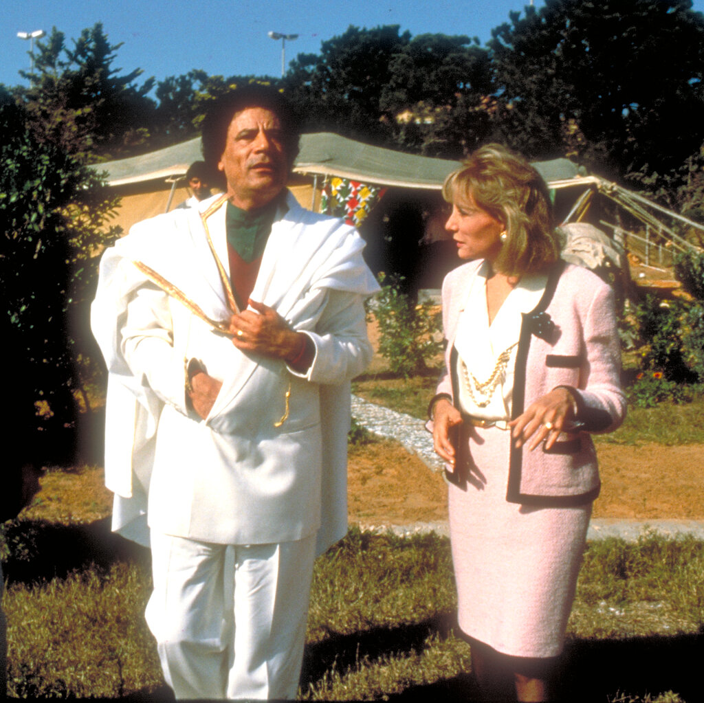 A TV still showing Col. Muammar el-Qaddafi, in a white suit, walking in a grassy field with Barbara Walters, who is wearing a pink skirt suit and pearls.