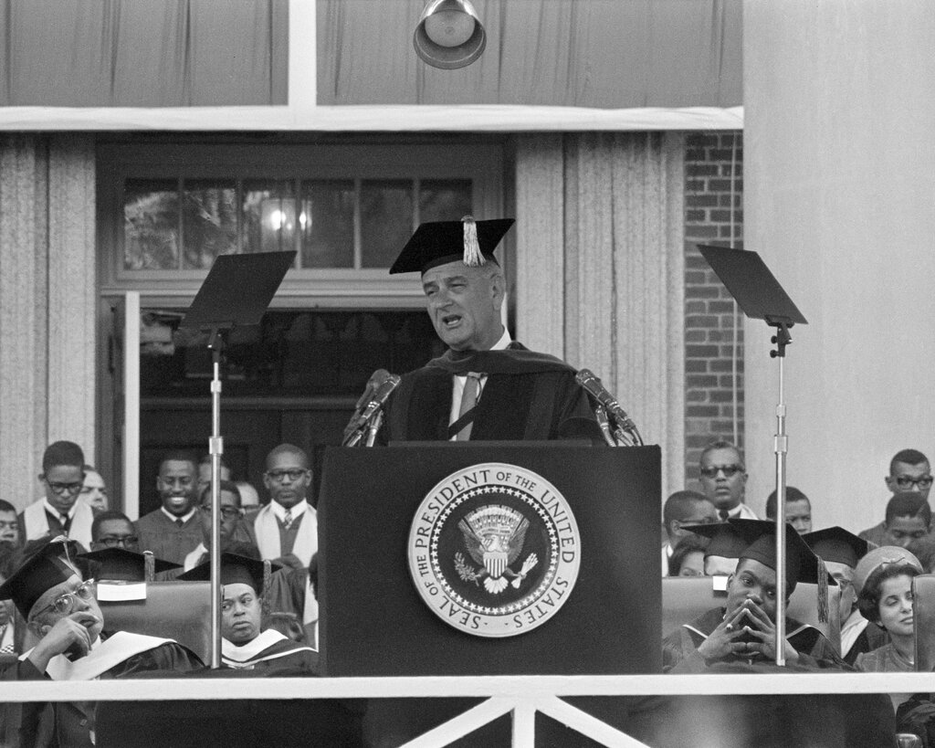 In a black-and-white photo, President Lyndon Johnson, wearing an academic gown and mortar board, delivers a speech at a podium.