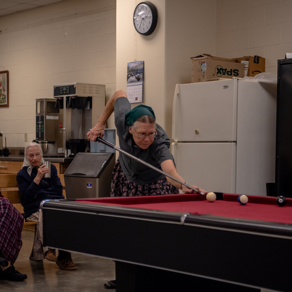 An older woman, leaning over a pool table, draws back a cue. 