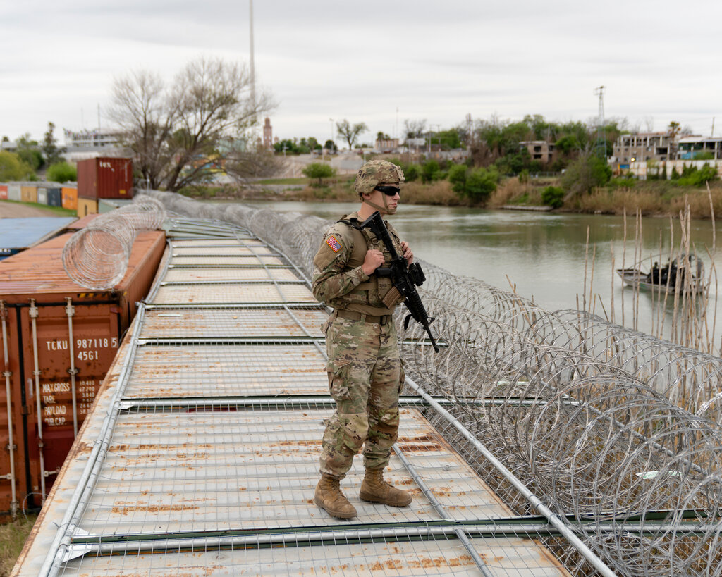 A U.S. National Guardsman, wearing fatigues and sunglasses and holding a rifle, stands on shipping containers with barbed wire at a riverbank.