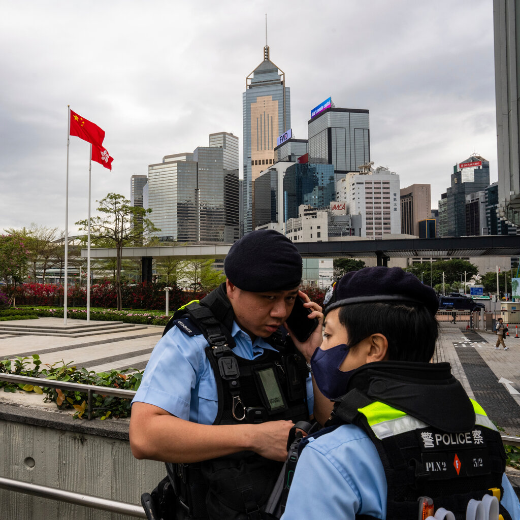 Uniformed police officers stand near a railing, with one using a cellphone. The flags of China and Hong Kong wave in a nearby plaza.