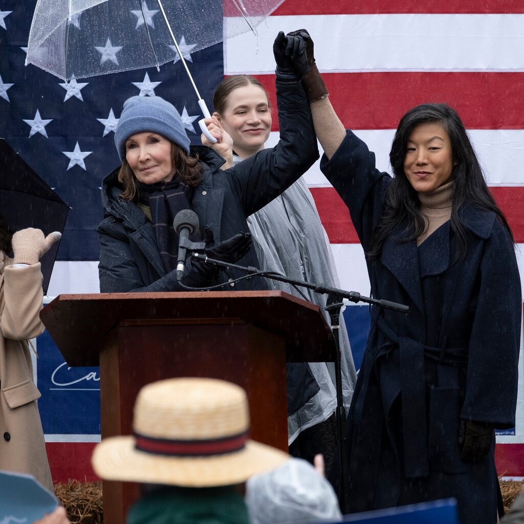 Two women stand at a podium in front of an American flag, their hands joined and held in the air. 