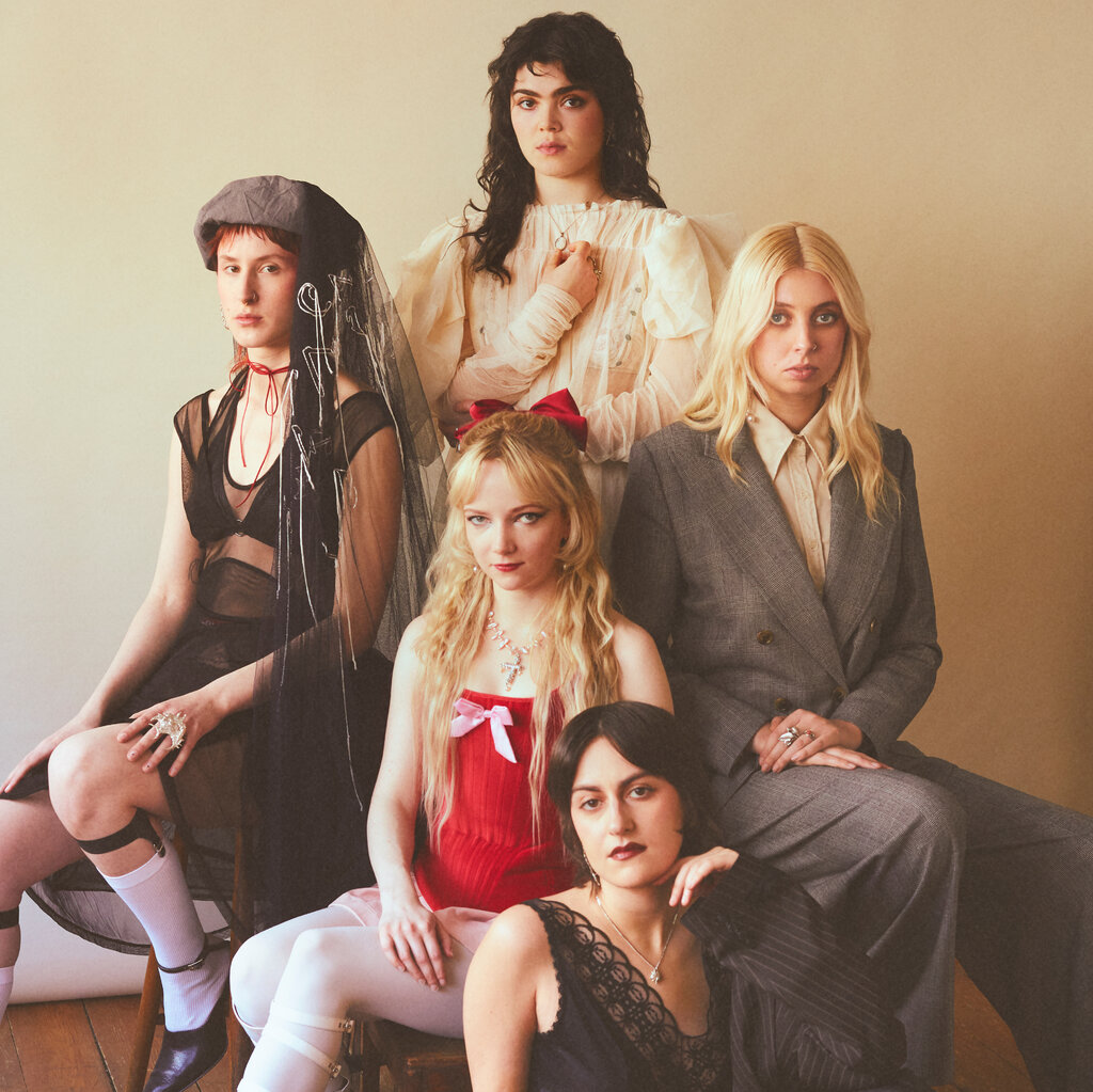 Five people sit and stand, wearing a mix of masculine and feminine outfits, all looking directly at the camera.