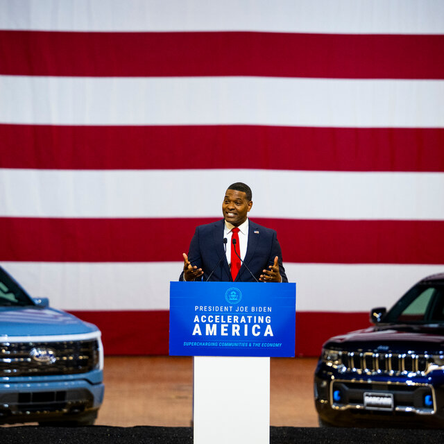 A man in a blue business suit and red tie stands at a podium and is flanked on either side by a shiny new electric vehicle. The backdrop is an enormous American flag. 