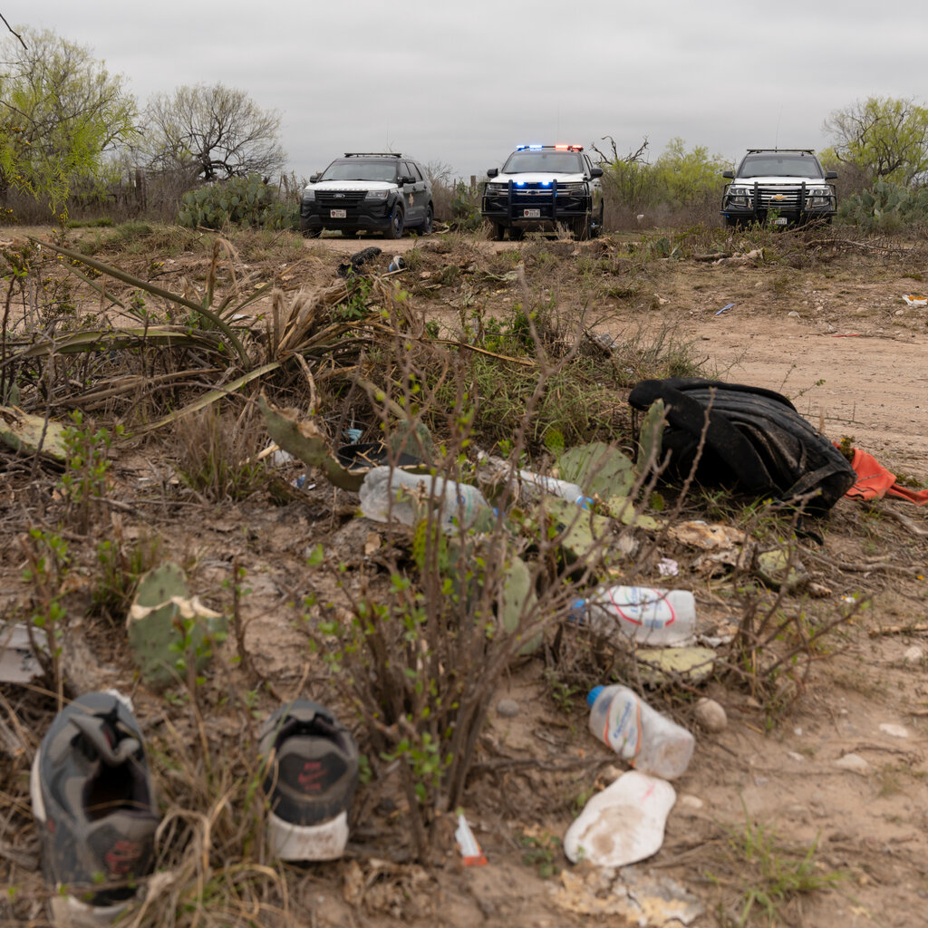 Discarded water bottles, shoes and a backpack are visible in dusty shrubbery, with three police vehicles in the background.