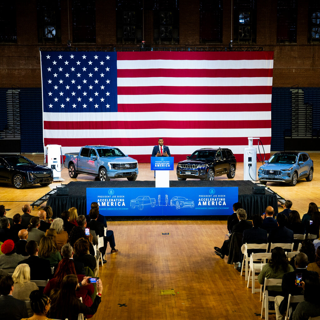 A man in a suit stands onstage with cars in front of a large American flag. 