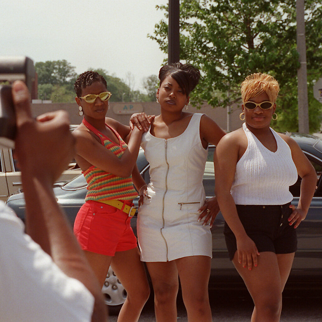 Three Black women pose for a camera held by a man in the foreground of the photo. The woman on the left sports finger waves and the women to her right both wear short, high-volume hairdos.