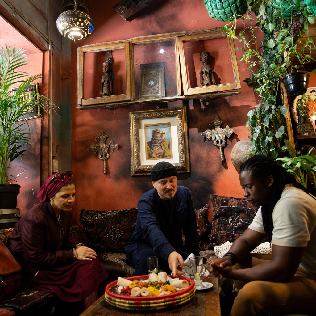 Three people sit around a table of food against a red wall with plants and African art in frames. 