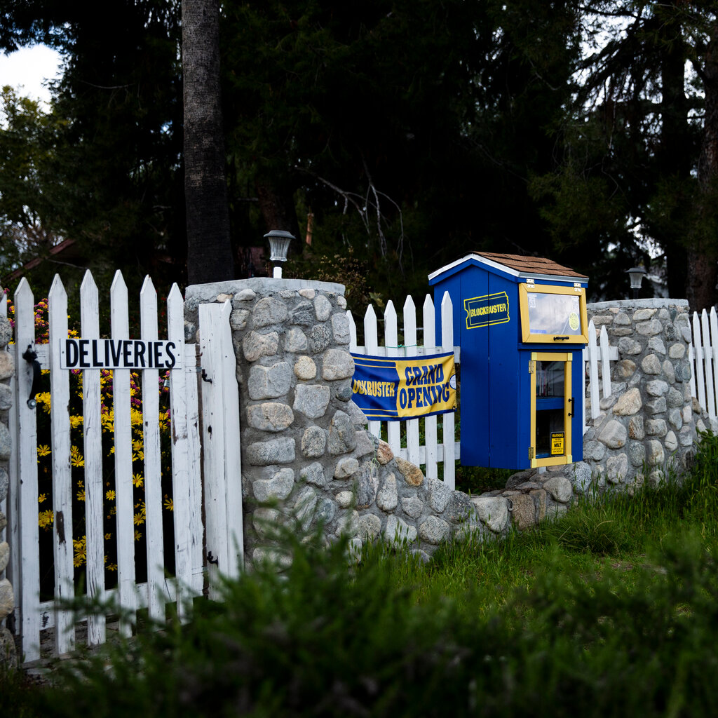 A miniature library, painted blue and yellow with the Blockbuster logo and a sign saying “Grand Opening,” sits on a white fence.