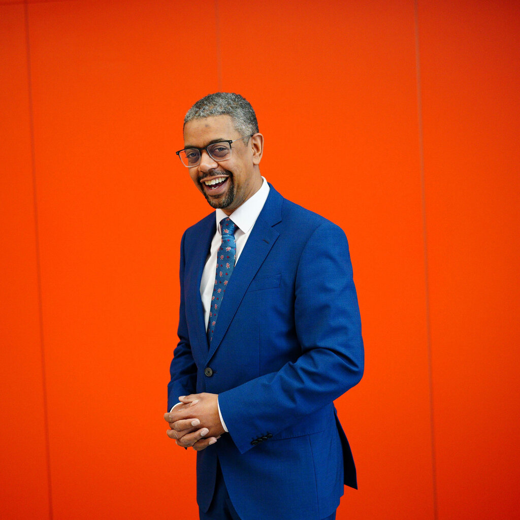 A man in glasses and a blue suit in front of a red wall. 