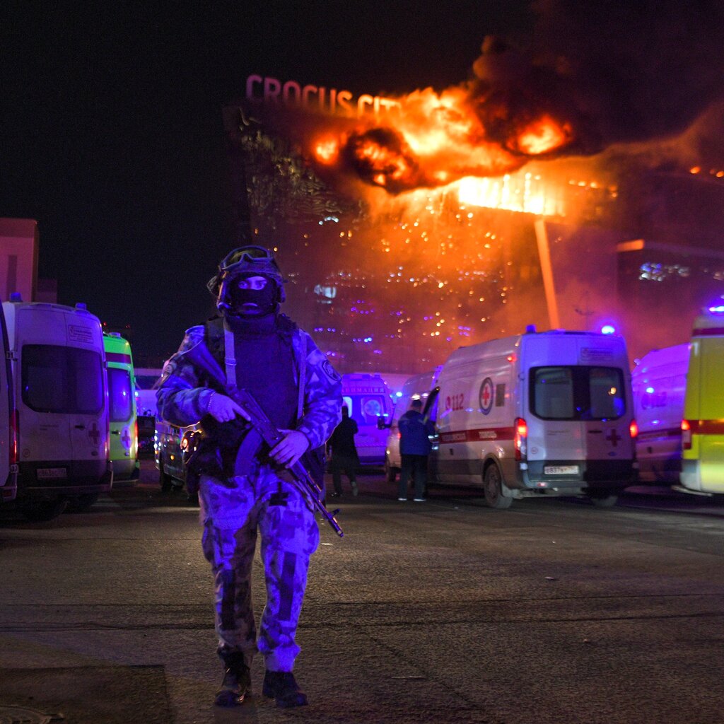 A man in a military uniform with a rife walking in a parking lot with a burning building behind him.