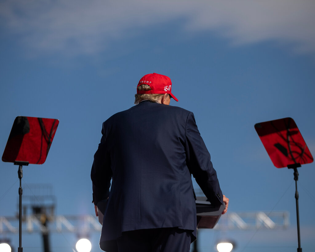 Former President Donald Trump is pictured from behind. He is standing at a lectern and wearing a red cap. 
