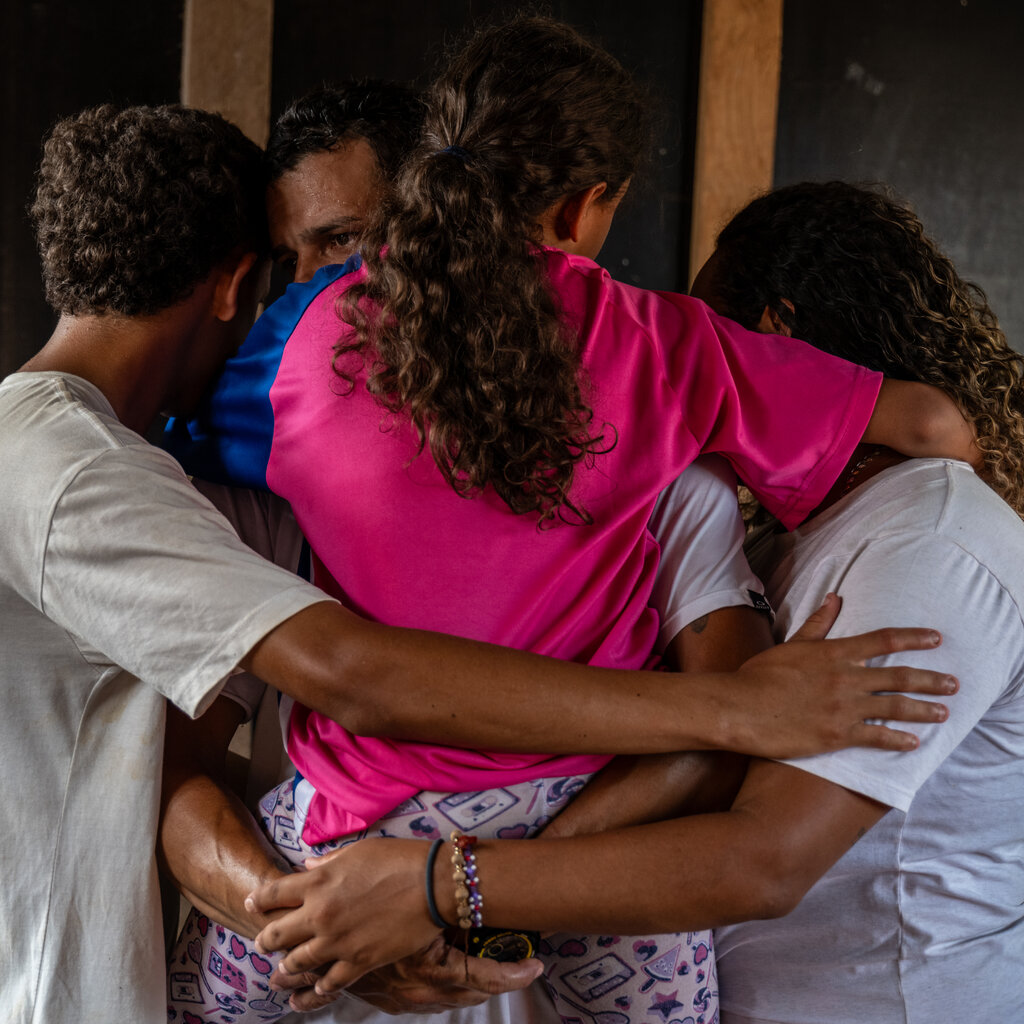A group of four people, including a child in a pink shirt, embrace in a group hug.