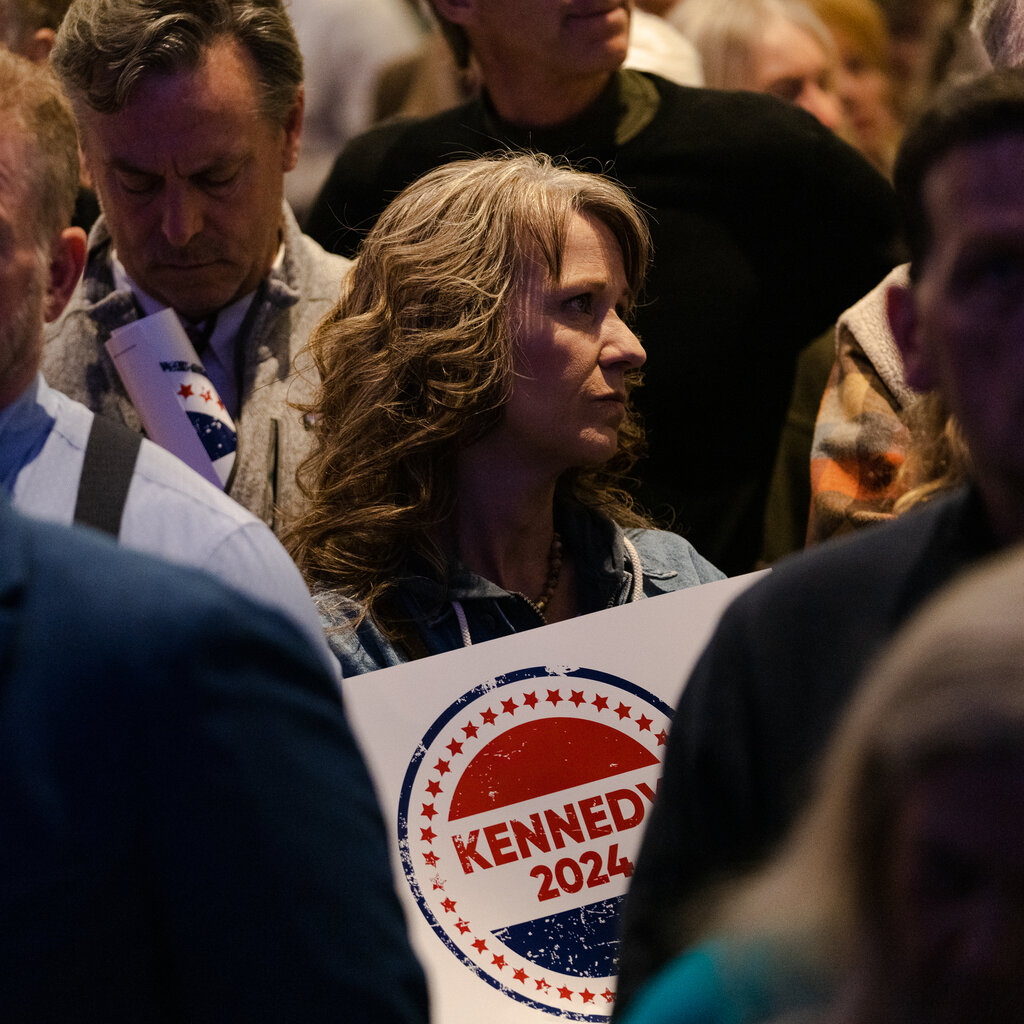 A woman stands in a crowd, a “Kennedy 2024” campaign sign hanging around her neck.