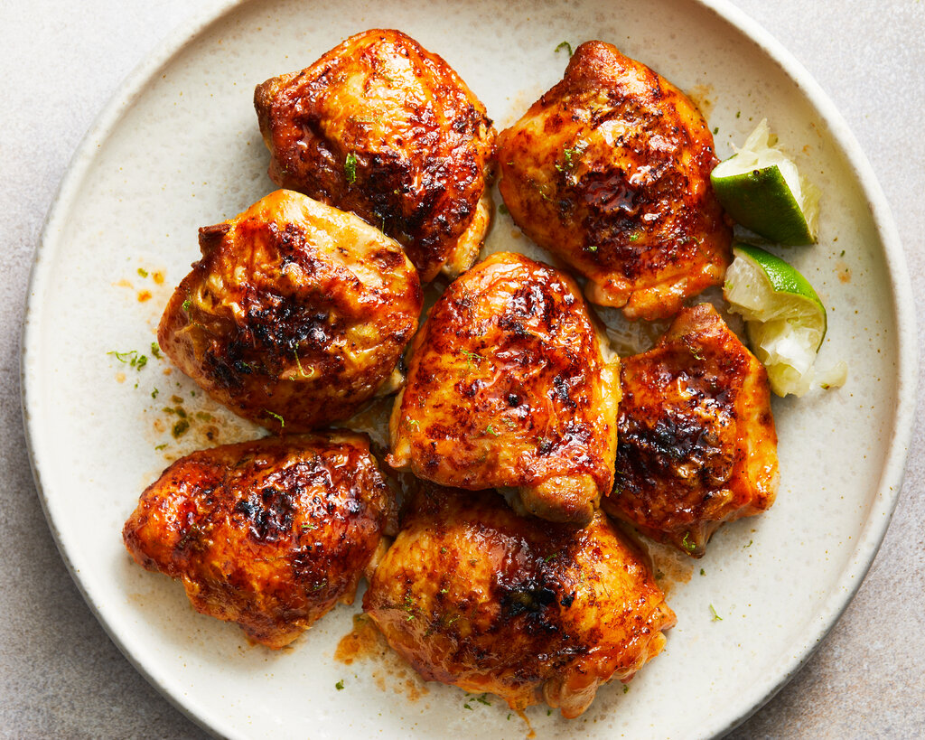 Seven roasted chicken thighs with hot honey and lime are on an ivory plate with squeezed lime wedges.