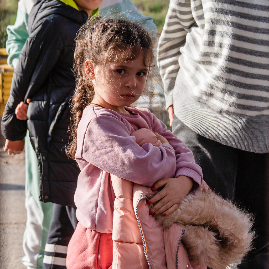A young girl in a pink outfit and braids holds her coat in an outdoor area.