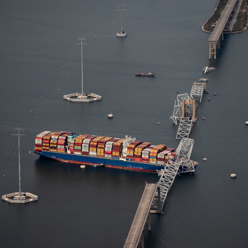 An aerial view of a cargo ship in a body of water. A portion of a bridge is collapsed on top of it. 