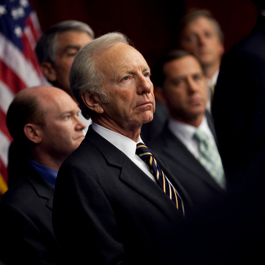 Senator Joe Lieberman, a formally dressed man with white hair, stands with other similarly dressed men in front of an American flag.