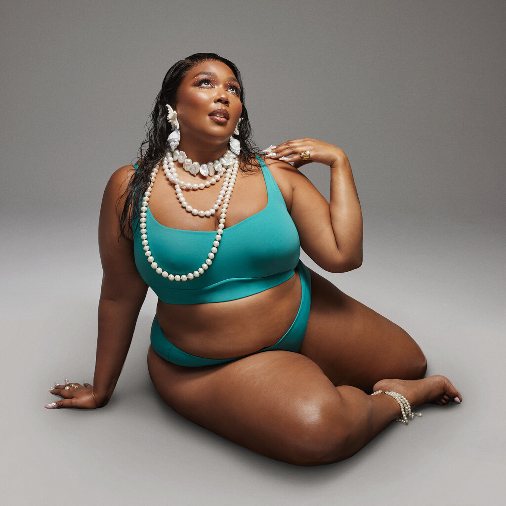 Lizzo posing in front of a gray backdrop in a teal two-piece swimsuit. She has long dark hair styled in wet waves, and she is wearing shell-shaped earrings and several pearl necklaces.