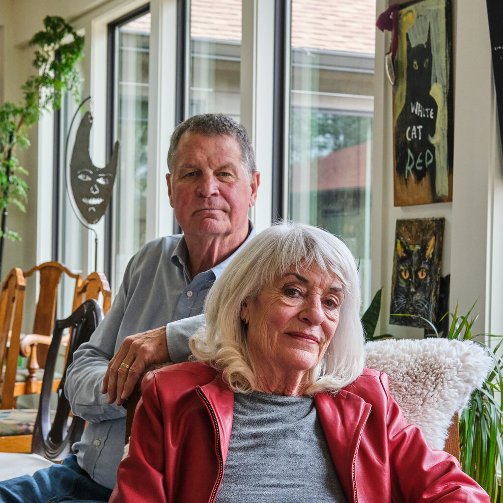 An older couple sit in their home with a dining room table behind them and colorful paintings on the wall.
