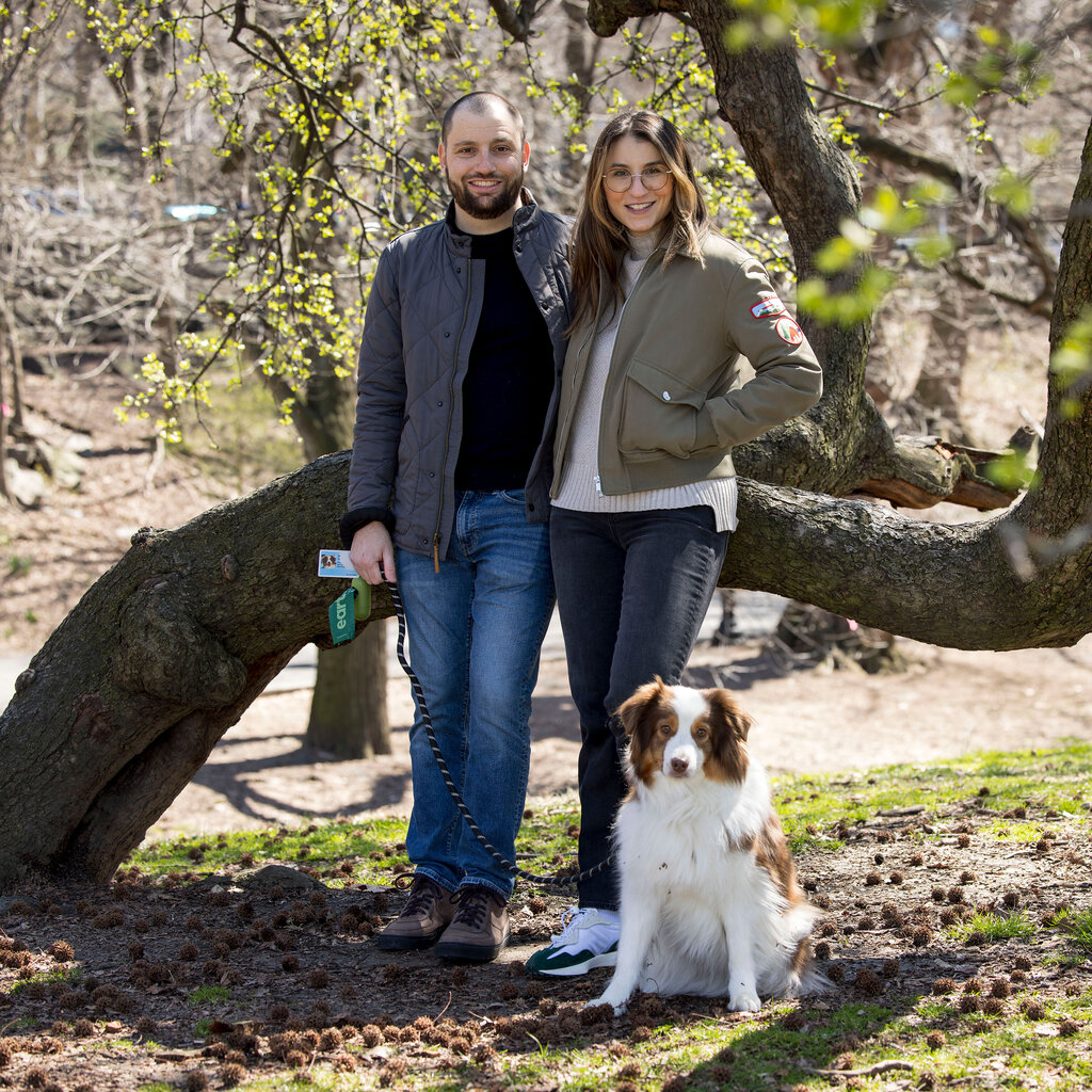 A couple stands with a dog in front of a tree in a park. 