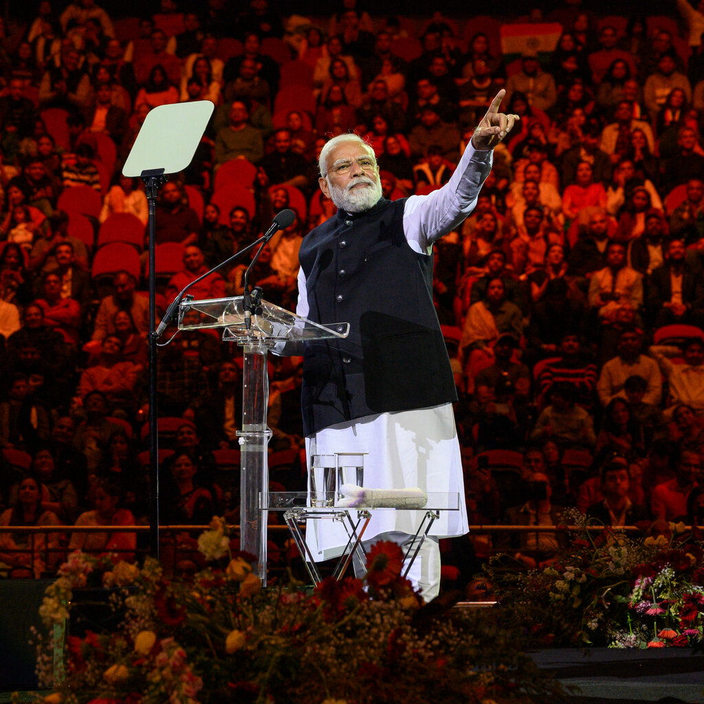 Narendra Modi in a long white shirt and a black vest stands onstage and points up. A crowd is visible behind him.