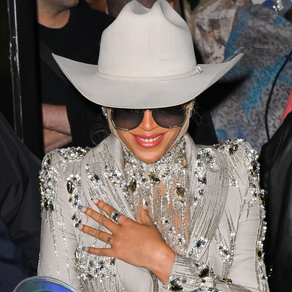 Beyoncé in a cowboy hat, sunglasses and elaborately bejeweled jacket brings one hand to her chest and smiles.