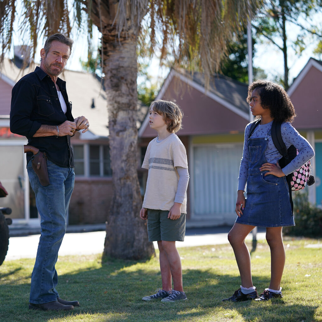 Dennis Quaid in bluejeans, with a gun on his hip, stands next to two children beneath a tree.