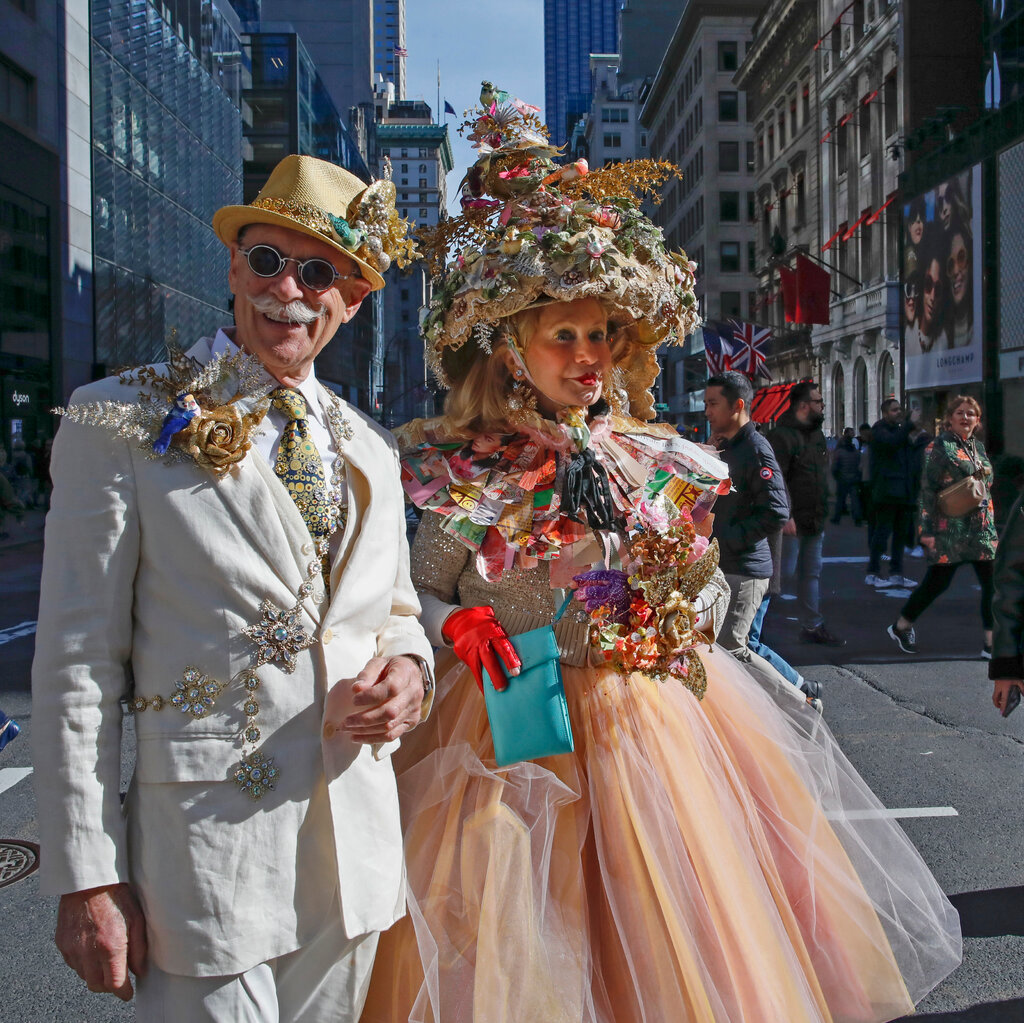 A man and woman walking down the street arm in arm. He has a mustache with curly ends and is wearing a light suit with a gold fedora and various sparkling accessories. She is wearing a dress with a puffy, peach-pink skirt and an elaborate, multicolor collar, and atop her head is a tall, pointed hat topped with a bird.