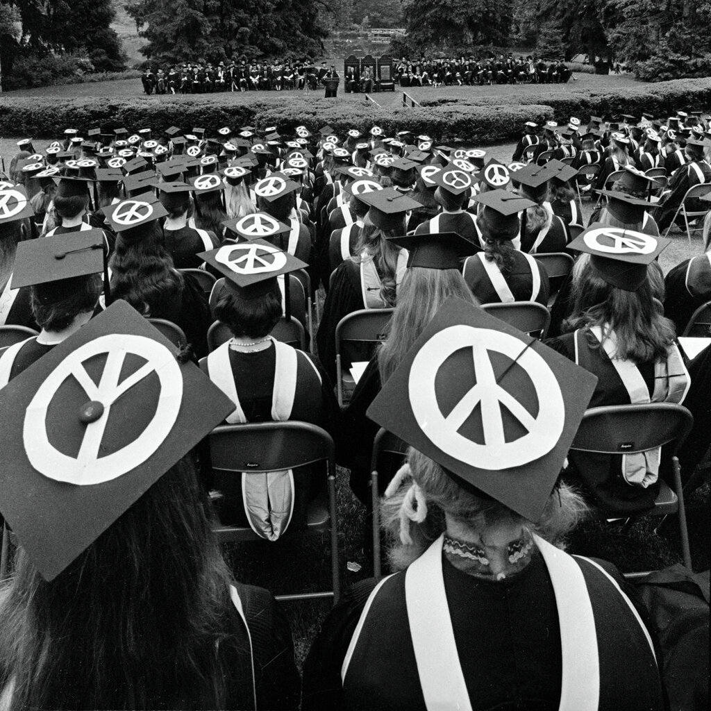 A black-and-white photo shows a sea of students at a graduation ceremony. They are wearing peace signs on their mortarboards.